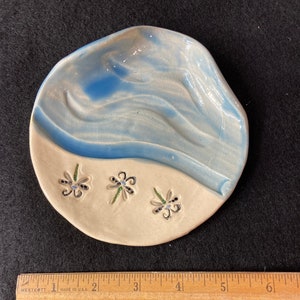 May include: A small, white ceramic dish with a blue glaze. The dish has a wavy design and three stylized flowers with black and green accents.
