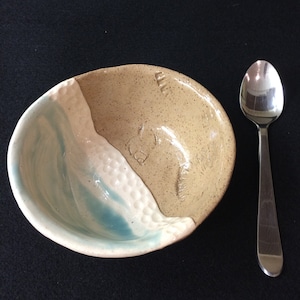 May include: A small, handmade ceramic bowl with a light blue and tan glaze. The bowl has a textured rim and a slightly uneven shape. It is sitting on a black surface next to a silver spoon.