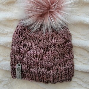 May include: A knitted beanie with a pink and white pom pom. The beanie is a deep mauve color and has a cable knit pattern. The beanie has a small metal tag that says "Eulalie Kiss Knits".