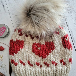Puede incluir: Patrón de tejido para un gorro blanco y rojo con un pompón de piel sintética. El gorro tiene un patrón de corazones.
