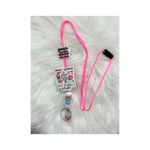 May include: Pink lanyard with a white charm that says "Busy Doing Cool Teacher Stuff" with a variety of school supplies on it. The charm is attached to a white charm that says "Super Teacher" with a stack of pencils on it. The lanyard has a clear badge holder.