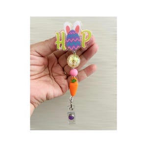 May include: A colourful Easter-themed badge reel featuring a purple and pink egg, bunny ears, and the word "HOP" in green letters. The reel has a pink and yellow bead, a carrot charm, and a clear badge clip with a purple button.