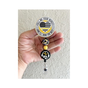 May include: A retractable badge reel with a round design. The top design features a yellow and black American flag with the text "TELL ME YOUR WORST AND I'LL SEND THE BEST". Below are decorative beads and a black 911 design.