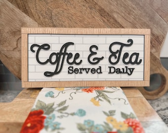 May include: A wooden sign with a white brick background and black lettering that reads "Coffee & Tea Served Daily".