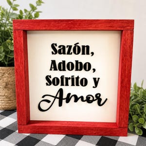 May include: A red framed wooden sign with the words "Sazón, Adobo, Sofrito y Amor" in black script. The sign is on a white background and is surrounded by greenery.