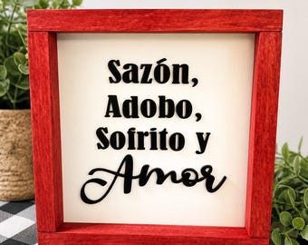 May include: A red framed wooden sign with the words "Sazón, Adobo, Sofrito y Amor" in black script. The sign is on a white background and is surrounded by greenery.