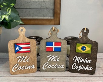 May include: Three wooden cutting boards with flags and the words "Mi Cocina" or "Minha Cozinha" painted on them. The flags are the Puerto Rican flag, the Dominican Republic flag, and the Brazilian flag.