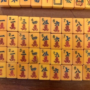 May include: A collection of vintage Mahjong tiles, featuring a warm yellow hue with red Chinese characters. The tiles are arranged in rows, showcasing various numbers and symbols, perfect for game enthusiasts.