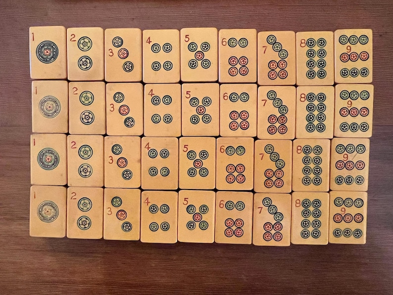 K&ouml;nnte beinhalten: Eine Sammlung von Vintage-Mahjong-Steinen, jeder mit einzigartigen Markierungen und Zahlen. Die Steine sind beige mit roten, gr&uuml;nen und blauen Symbolen. Die Zahlen 1 bis 9 sind auf den Steinen abgebildet, zusammen mit verschiedenen Symbolen.