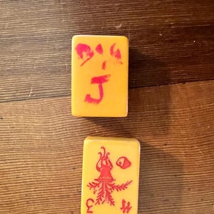 May include: Two rectangular, yellow Mahjong tiles with red symbols. One tile has a large red "J" and other symbols. The second tile has a red illustration of a figure, a "3", and other symbols. The tiles are on a wooden surface.