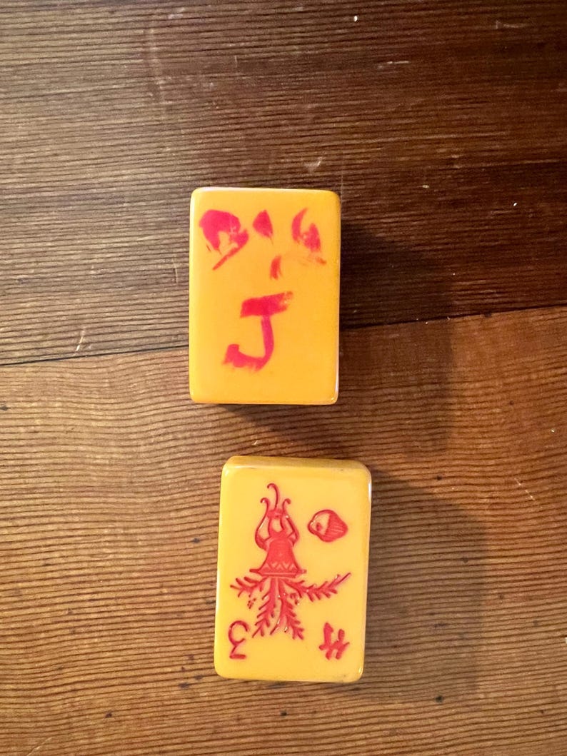 May include: Two rectangular, yellow Mahjong tiles with red symbols. One tile has a large red "J" and other symbols. The second tile has a red illustration of a figure, a "3", and other symbols. The tiles are on a wooden surface.