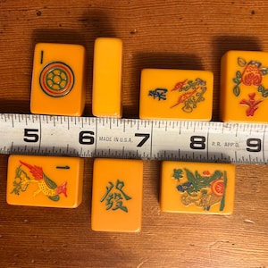 May include: A collection of vintage Mahjong tiles in shades of yellow and orange, featuring various symbols and characters. The tiles are arranged on a wooden surface, with a ruler providing scale. The tiles measure approximately 1.5 inches in length.