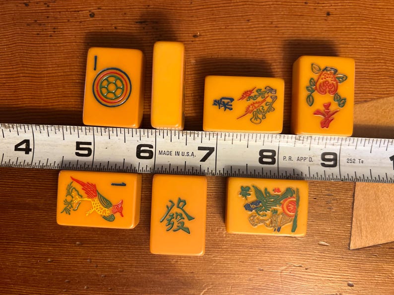 May include: A collection of vintage Mahjong tiles in shades of yellow and orange, featuring various symbols and characters. The tiles are arranged on a wooden surface, with a ruler providing scale. The tiles measure approximately 1.5 inches in length.