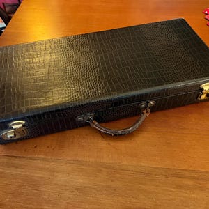 May include: A rectangular black case with a textured, crocodile-like pattern. The case has a brown handle and gold-colored latches. The case is on a wooden surface.