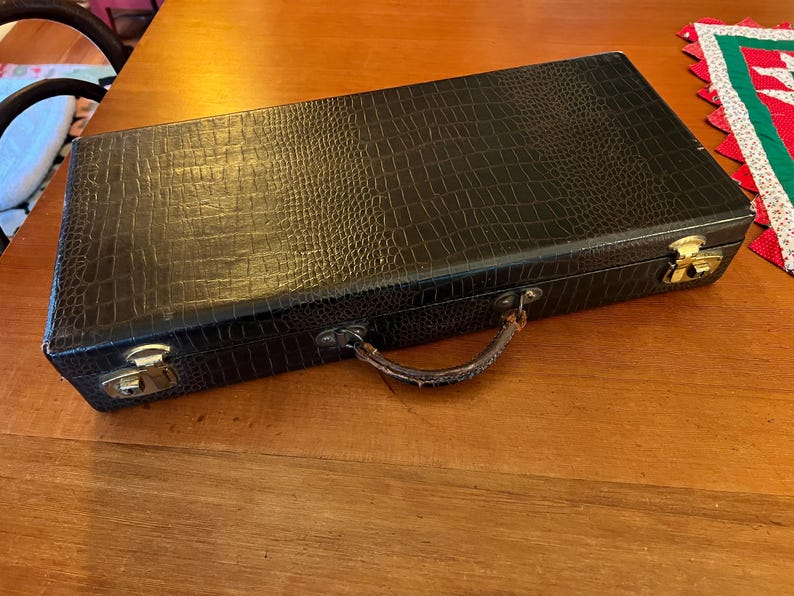 May include: A rectangular black case with a textured, crocodile-like pattern. The case has a brown handle and gold-colored latches. The case is on a wooden surface.