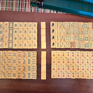 K&ouml;nnte beinhalten: Ein komplettes Mahjong-Set mit elfenbeinfarbenen Spielsteinen, angeordnet auf einer Holzoberfl&auml;che. Die Spielsteine zeigen chinesische Schriftzeichen, Symbole und Illustrationen. Das Set enth&auml;lt eine Vielzahl von Spielsteinen.