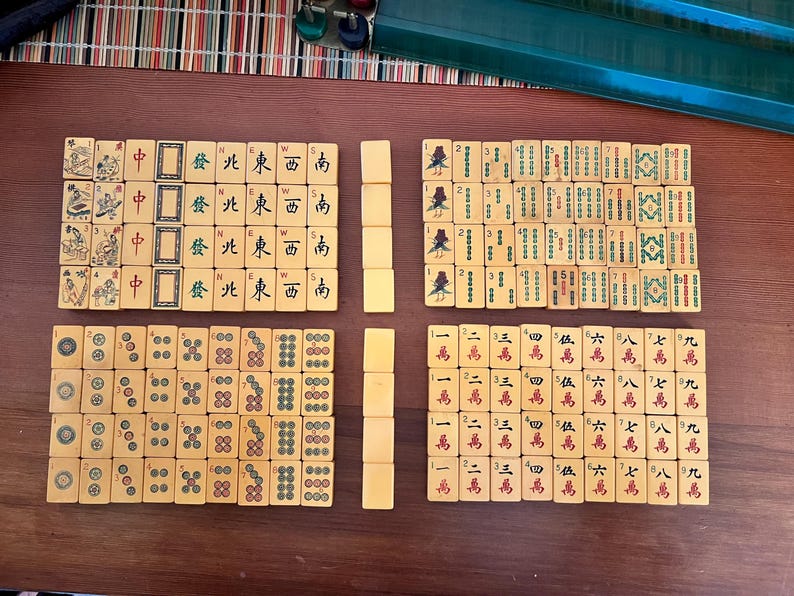 K&ouml;nnte beinhalten: Ein komplettes Mahjong-Set mit elfenbeinfarbenen Spielsteinen, angeordnet auf einer Holzoberfl&auml;che. Die Spielsteine zeigen chinesische Schriftzeichen, Symbole und Illustrationen. Das Set enth&auml;lt eine Vielzahl von Spielsteinen.