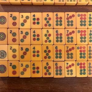 May include: A collection of vintage Mahjong tiles, featuring ivory-colored tiles with red, green, and blue markings. The tiles display numbers and suits, arranged on a wooden surface. The game pieces are in a grid pattern.