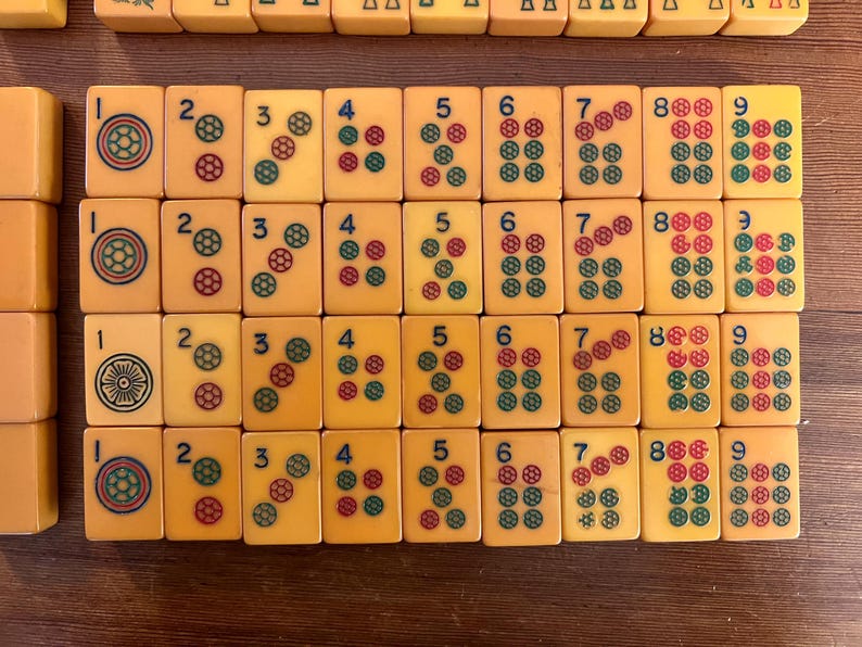 May include: A collection of vintage Mahjong tiles, featuring ivory-colored tiles with red, green, and blue markings. The tiles display numbers and suits, arranged on a wooden surface. The game pieces are in a grid pattern.