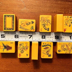 K&ouml;nnte beinhalten: Eine Sammlung gelber Vintage-Mahjong-Steine mit verschiedenen Illustrationen und Zahlen. Die Steine sind auf einer Holzoberfl&auml;che neben einem Lineal angeordnet. Die Steine sind etwa 2,5 cm breit.