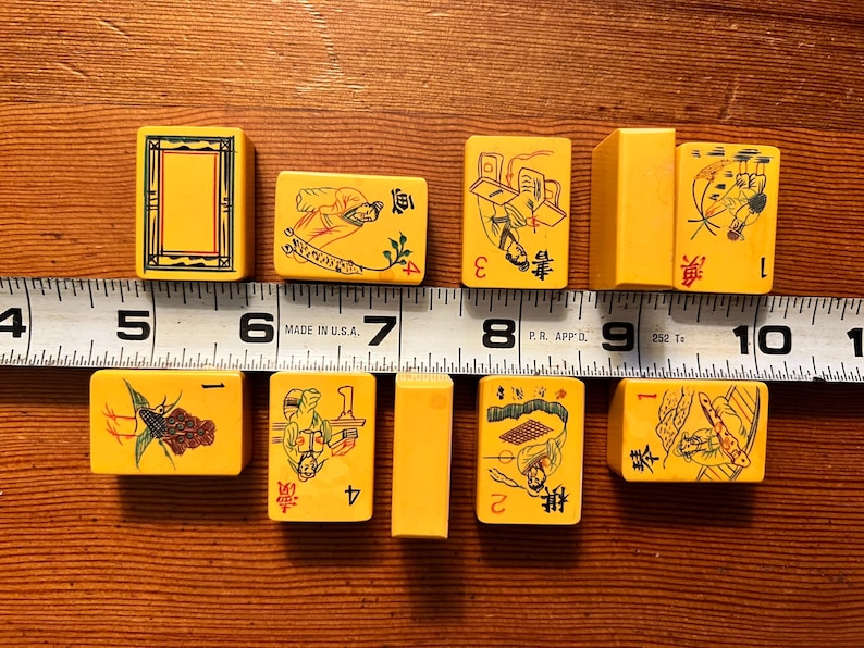 K&ouml;nnte beinhalten: Eine Sammlung gelber Vintage-Mahjong-Steine mit verschiedenen Illustrationen und Zahlen. Die Steine sind auf einer Holzoberfl&auml;che neben einem Lineal angeordnet. Die Steine sind etwa 2,5 cm breit.
