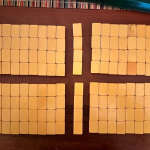 K&ouml;nnte beinhalten: Ein Satz Vintage-Mahjong-Steine, angeordnet auf einer braunen Oberfl&auml;che. Die rechteckigen Steine sind hellgelb. Das Layout deutet auf ein laufendes Spiel hin, mit in S&auml;tzen gruppierten Steinen.