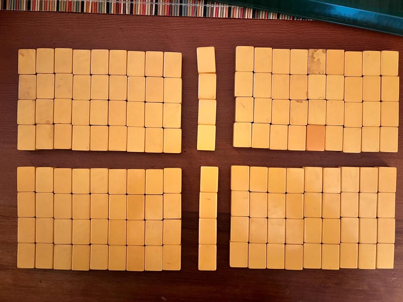 K&ouml;nnte beinhalten: Ein Satz Vintage-Mahjong-Steine, angeordnet auf einer braunen Oberfl&auml;che. Die rechteckigen Steine sind hellgelb. Das Layout deutet auf ein laufendes Spiel hin, mit in S&auml;tzen gruppierten Steinen.