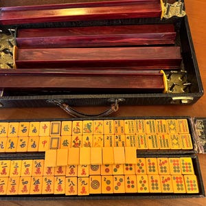 May include: A vintage Mahjong set in a black case with a handle. The set includes wooden racks and tiles with Chinese characters and symbols. The tiles are a light orange color.