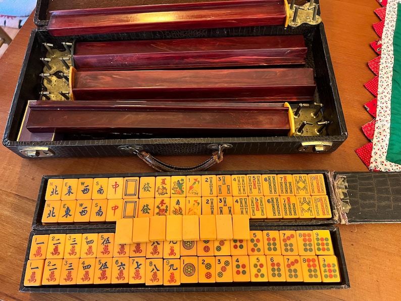 May include: A vintage Mahjong set in a black case with a handle. The set includes wooden racks and tiles with Chinese characters and symbols. The tiles are a light orange color.