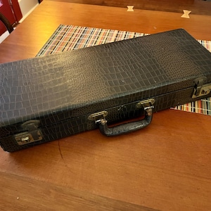 May include: A dark gray, rectangular case with a textured, crocodile-like pattern. The case has a handle and metal clasps. It is resting on a wooden table with a colorful striped placemat.