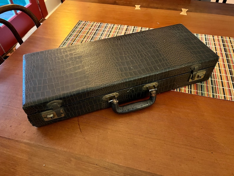 K&ouml;nnte beinhalten: Ein dunkelgrauer, rechteckiger Koffer mit einem strukturierten, krokodilartigen Muster. Der Koffer hat einen Griff und Metallverschl&uuml;sse. Er liegt auf einem Holztisch mit einem bunten gestreiften Tischset.