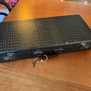 Puede incluir: Un estuche rectangular negro con textura de piel de cocodrilo sintética. El estuche tiene un asa marrón, cierres metálicos y una llave sujeta con una cuerda blanca. El estuche está cerrado y descansa sobre una superficie de madera.