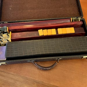 May include: Open vintage Mahjong set in a dark brown case with a handle. The case contains wooden game pieces, racks, and tiles. The tiles are yellow and rectangular. The case is open on a wooden surface.