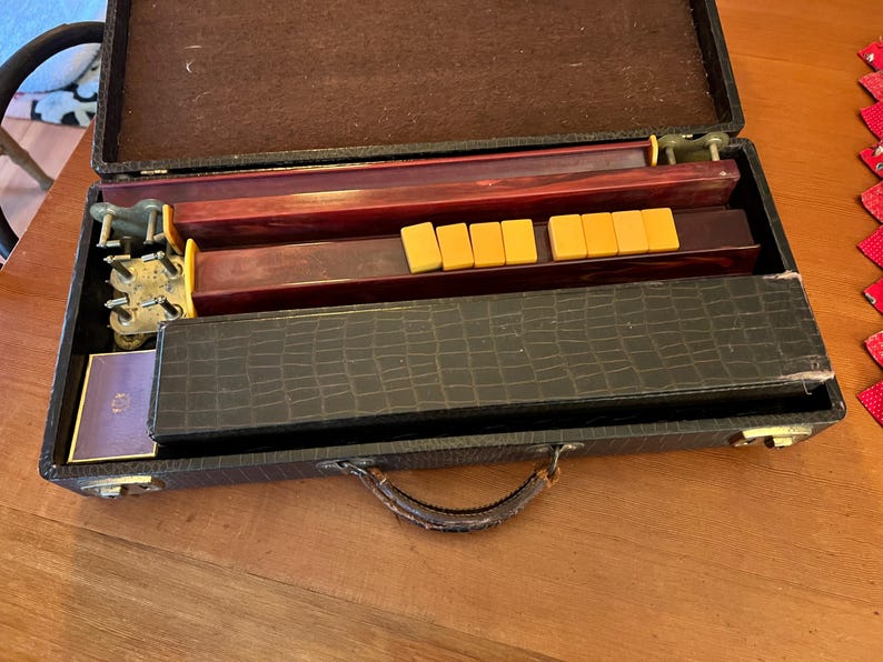 May include: Open vintage Mahjong set in a dark brown case with a handle. The case contains wooden game pieces, racks, and tiles. The tiles are yellow and rectangular. The case is open on a wooden surface.