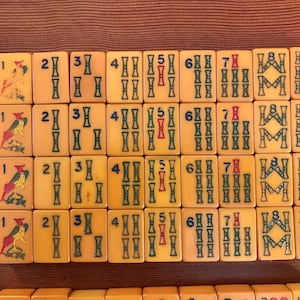 May include: A collection of vintage Mahjong tiles, arranged in rows. The tiles are a light yellow color with green, red, and blue markings. Some tiles feature bird illustrations. The numbers and symbols are clearly visible.