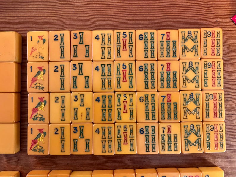 May include: A collection of vintage Mahjong tiles, arranged in rows. The tiles are a light yellow color with green, red, and blue markings. Some tiles feature bird illustrations. The numbers and symbols are clearly visible.