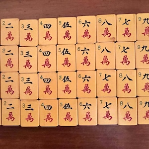 K&ouml;nnte beinhalten: Eine Sammlung von Vintage-Mahjong-Steinen auf einer Holzoberfl&auml;che. Die cremefarbenen Steine ​​sind mit roten chinesischen Schriftzeichen und Zahlen versehen. Die Spielsteine ​​sind in Reihen und Spalten angeordnet.