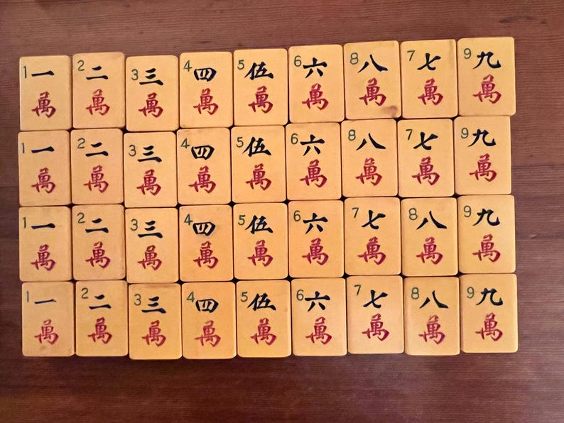 K&ouml;nnte beinhalten: Eine Sammlung von Vintage-Mahjong-Steinen auf einer Holzoberfl&auml;che. Die cremefarbenen Steine ​​sind mit roten chinesischen Schriftzeichen und Zahlen versehen. Die Spielsteine ​​sind in Reihen und Spalten angeordnet.