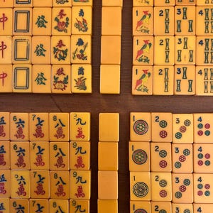 May include: A collection of vintage Mahjong tiles, featuring ivory-colored tiles with red, green, and blue symbols and characters. The tiles are arranged in several groups, showcasing various suits and numbers.
