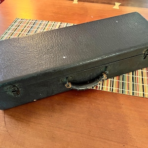 May include: A rectangular black case with a textured surface, metal clasps, and a handle. The case is resting on a colourful striped mat on a wooden table. The case appears to be for a musical instrument.