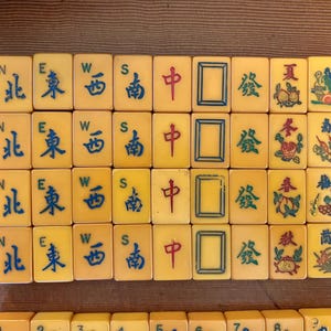 May include: A collection of vintage Mahjong tiles, featuring yellow tiles with blue and red Chinese characters and symbols. The tiles are arranged in rows, showcasing various suits and directions.