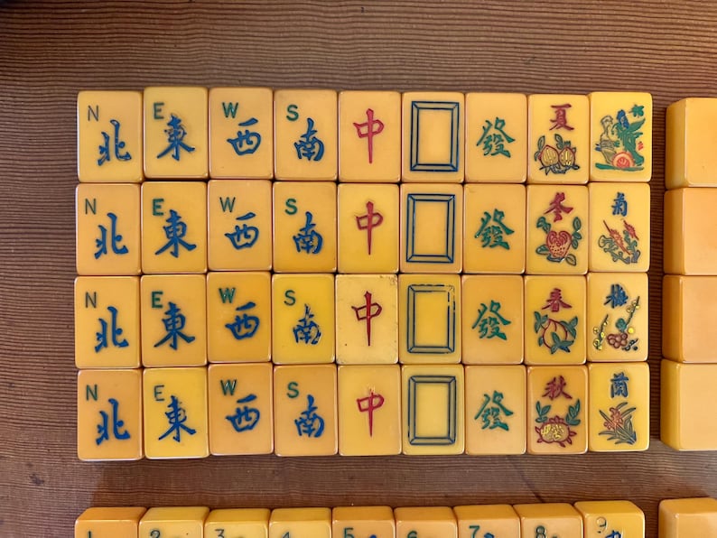May include: A collection of vintage Mahjong tiles, featuring yellow tiles with blue and red Chinese characters and symbols. The tiles are arranged in rows, showcasing various suits and directions.