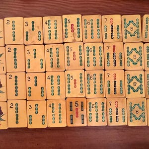 K&ouml;nnte beinhalten: Ein Satz Vintage-Mahjong-Steine, angeordnet auf einer Holzoberfl&auml;che. Die Steine sind cremefarben mit gr&uuml;nen und roten Markierungen und zeigen Zahlen, Symbole und dekorative Motive. Das Set enth&auml;lt Steine mit den Nummern 1 bis 9.