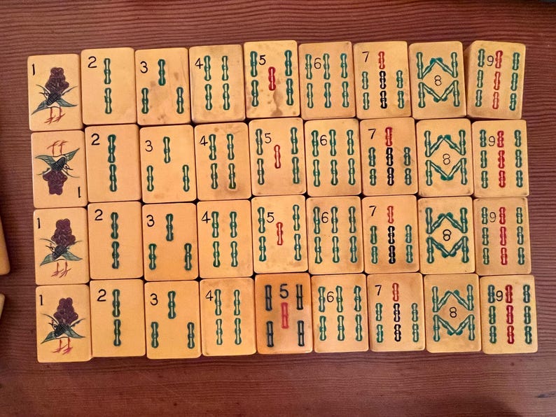 K&ouml;nnte beinhalten: Ein Satz Vintage-Mahjong-Steine, angeordnet auf einer Holzoberfl&auml;che. Die Steine sind cremefarben mit gr&uuml;nen und roten Markierungen und zeigen Zahlen, Symbole und dekorative Motive. Das Set enth&auml;lt Steine mit den Nummern 1 bis 9.