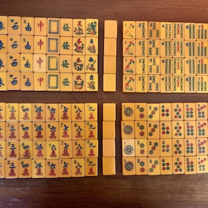 May include: A complete set of vintage Mahjong tiles, arranged on a wooden surface. The tiles are cream-colored with black, red, and green markings, featuring Chinese characters and symbols. The set includes tiles with numbers, suits, and wind directions.