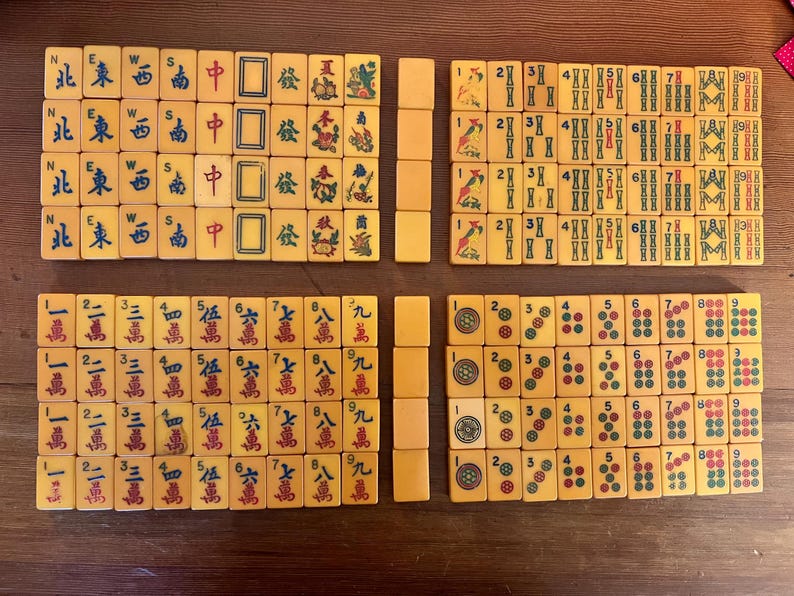 May include: A complete set of vintage Mahjong tiles, arranged on a wooden surface. The tiles are cream-colored with black, red, and green markings, featuring Chinese characters and symbols. The set includes tiles with numbers, suits, and wind directions.