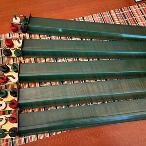 K&ouml;nnte beinhalten: Ein Set aus sechs gr&uuml;nen Mahjong-Kartenhaltern mit Messingfarbenen Haltern. Jeder Halter hat eine lange, rechteckige Form. Die Halter haben rote, wei&szlig;e, schwarze und gr&uuml;ne Kn&ouml;pfe. Die Halter sind auf einer gestreiften Matte angeordnet.