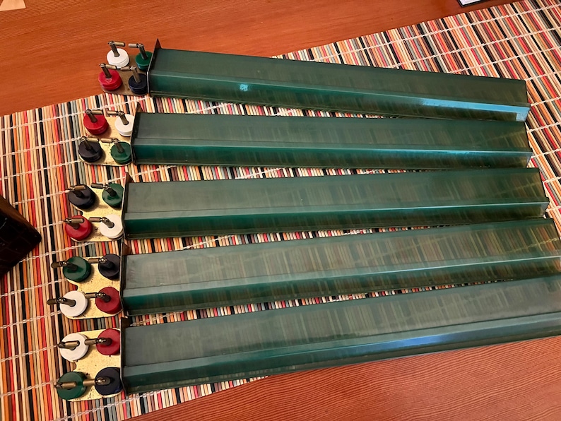 K&ouml;nnte beinhalten: Ein Set aus sechs gr&uuml;nen Mahjong-Kartenhaltern mit Messingfarbenen Haltern. Jeder Halter hat eine lange, rechteckige Form. Die Halter haben rote, wei&szlig;e, schwarze und gr&uuml;ne Kn&ouml;pfe. Die Halter sind auf einer gestreiften Matte angeordnet.