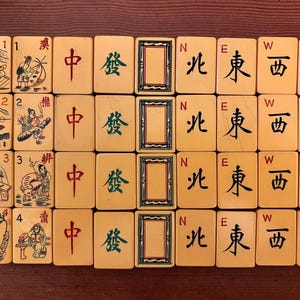 K&ouml;nnte beinhalten: Eine Sammlung von Vintage-Mahjong-Steinen, die auf einer Holzoberfl&auml;che angeordnet sind. Die Steine ​​weisen chinesische Schriftzeichen, Illustrationen und Richtungssymbole auf. Die Steine ​​sind cremefarben mit schwarzen, roten und gr&uuml;nen Markierungen und zeigen eine klassische Spiel&auml;sthetik.