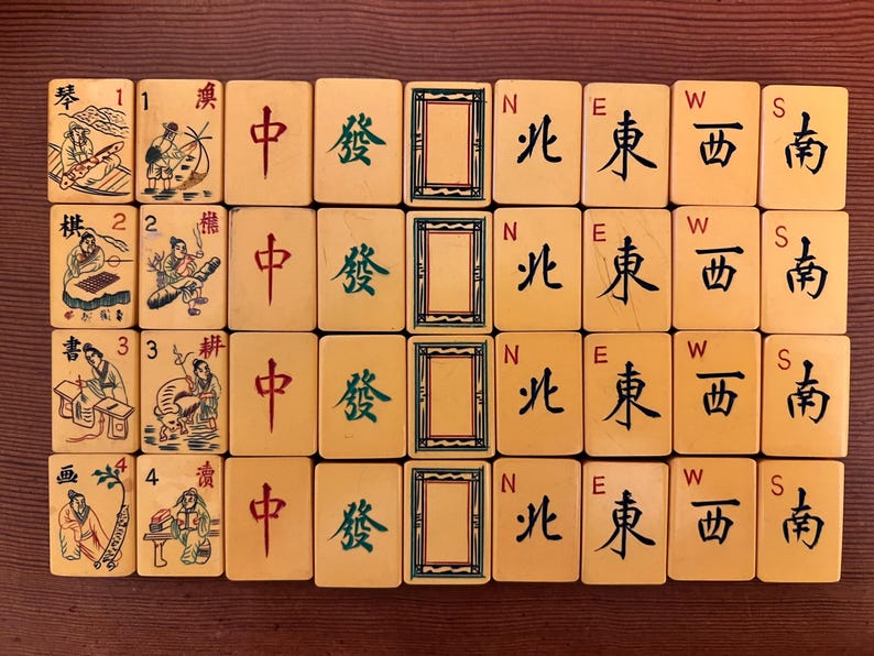 K&ouml;nnte beinhalten: Eine Sammlung von Vintage-Mahjong-Steinen, die auf einer Holzoberfl&auml;che angeordnet sind. Die Steine ​​weisen chinesische Schriftzeichen, Illustrationen und Richtungssymbole auf. Die Steine ​​sind cremefarben mit schwarzen, roten und gr&uuml;nen Markierungen und zeigen eine klassische Spiel&auml;sthetik.